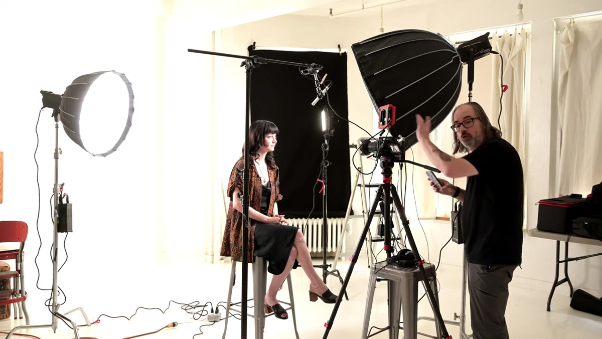 Interviewer stands at monitor beside camera, engaging subject